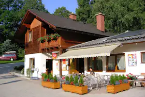 Holz-Ferienhaus mit Terrasse, Markise und vielen Stühlen, umgeben von Blumenbeeten und Bäumen im Campingbereich.