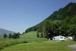Campingplatz am See mit Zelt und Wohnwagen am Ufer, grüner Rasen, Bäume und Berge im Hintergrund.