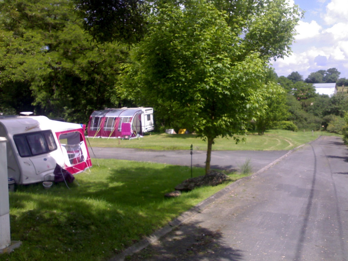 Wohnwagen und Zelt auf einer Campingwiese, daneben ein Auto, ein Baum und ein Weg entlang der Stellplätze.