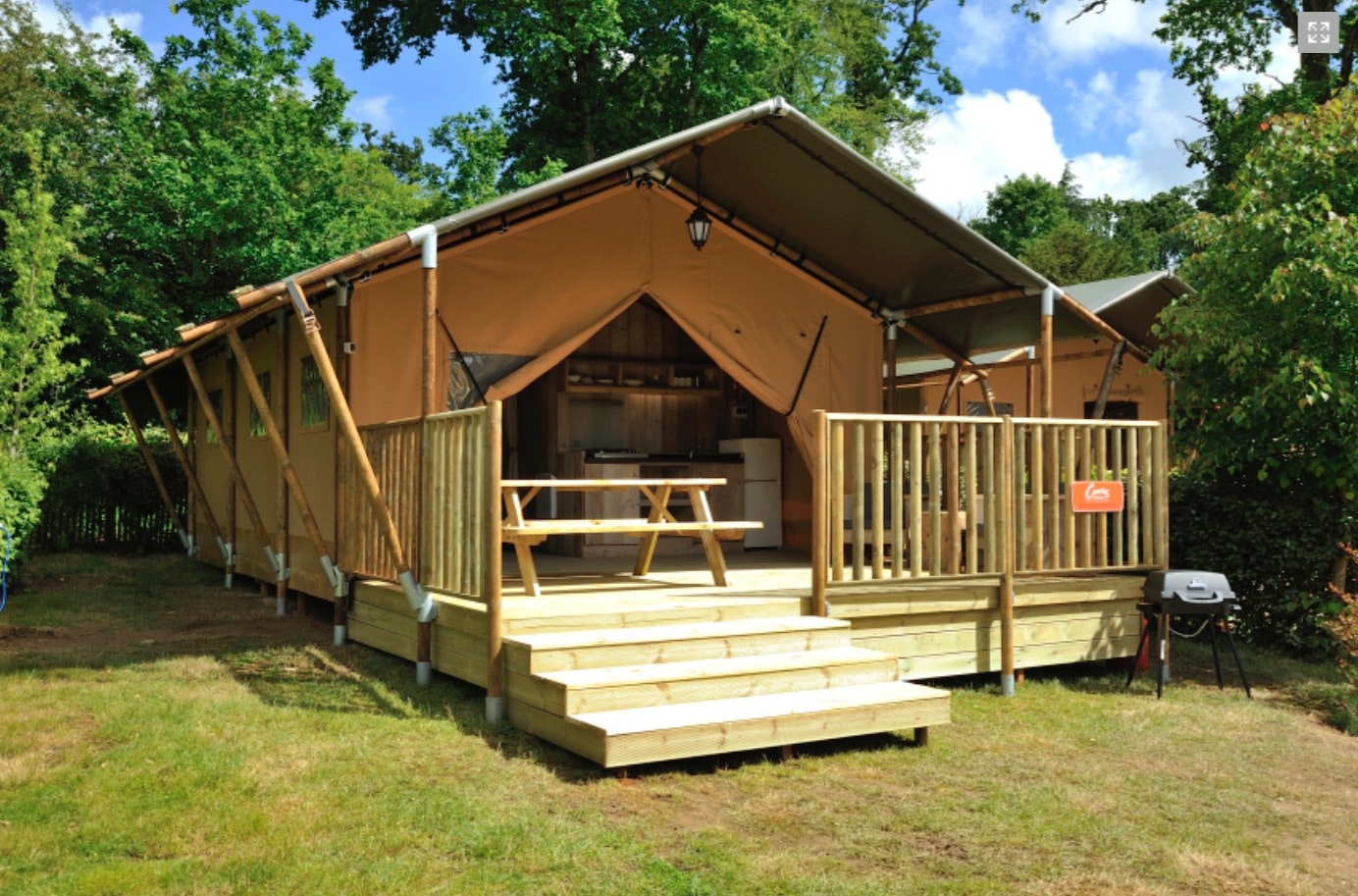 Zeltbungalow mit Holzveranda, Stufen und Sitzbank, umgeben von Bäumen und Rasen auf dem Campingplatz.