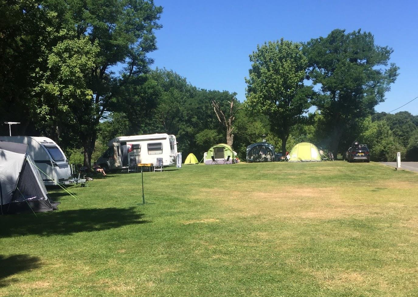 Campingplatz mit Zelten und Wohnwagen auf einer grünen Wiese, umgeben von Bäumen und einem Weg.