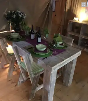 Dekorierter Camping-Tisch mit Geschirr, Weingläsern und zwei Weinflaschen im Zelt, daneben Blumenstrauß und Kerzenlicht.