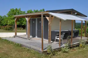 Camping-Unterkunft mit Holzveranda und Markise, daneben Tisch und Stühle, im Hintergrund Bäume und ein Hügel.
