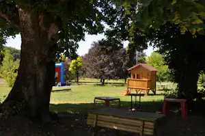 Holzspielhaus und Spielbereich auf dem Campingplatz im Grünen, mit Bank, Tisch und bunten Elementen unter einem großen Baum.