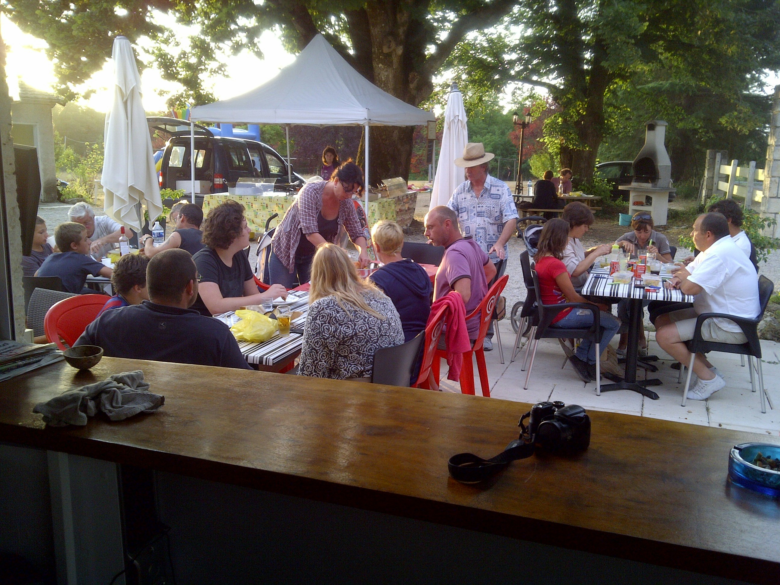 Gruppe am Camping-Tisch im Freien unter Bäumen, mit Sonnenschirmen, Autos im Hintergrund und Geschirr auf der Tischplatte.