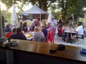 Gruppe am Camping-Tisch im Freien unter Bäumen, mit Sonnenschirmen, Autos im Hintergrund und Geschirr auf der Tischplatte.