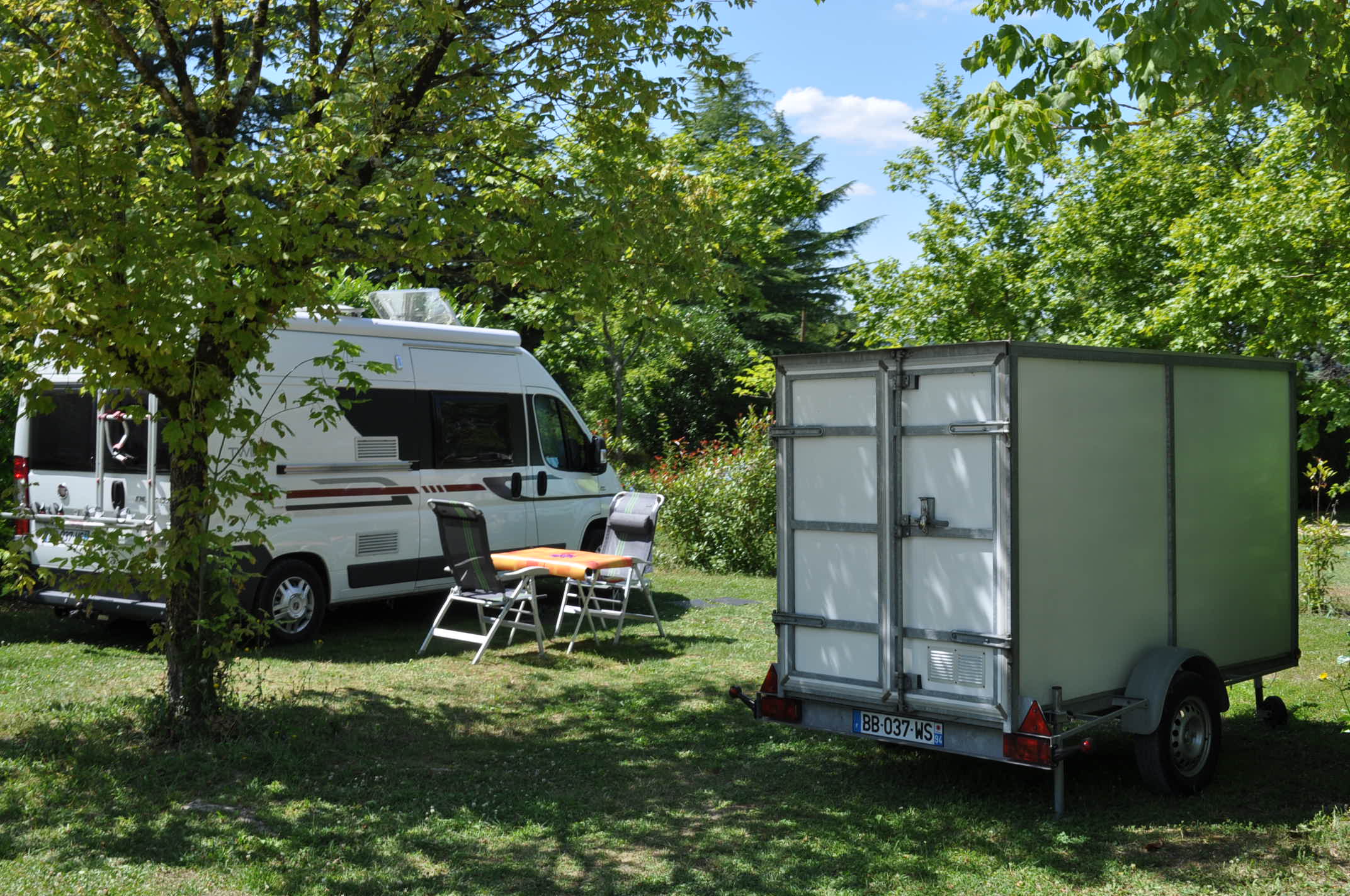 Wohnmobil und Anhänger auf einer Campingwiese, mit Gartenstühlen und Bäumen im Hintergrund.