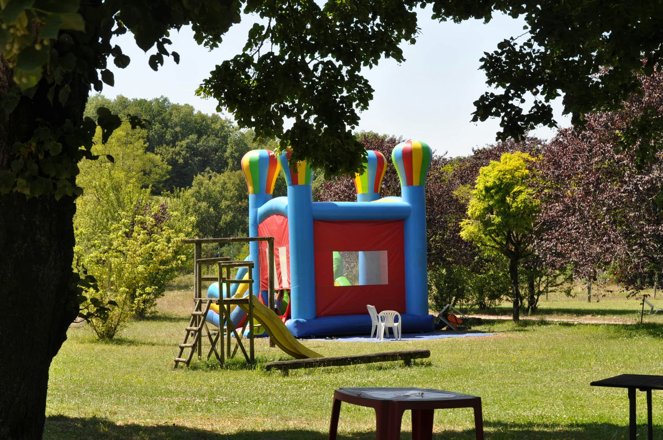 Aufblasbares Hüpfburg-Spielgerät mit Rutsche im grünen Campinggarten, umgeben von Bäumen und Wiese.