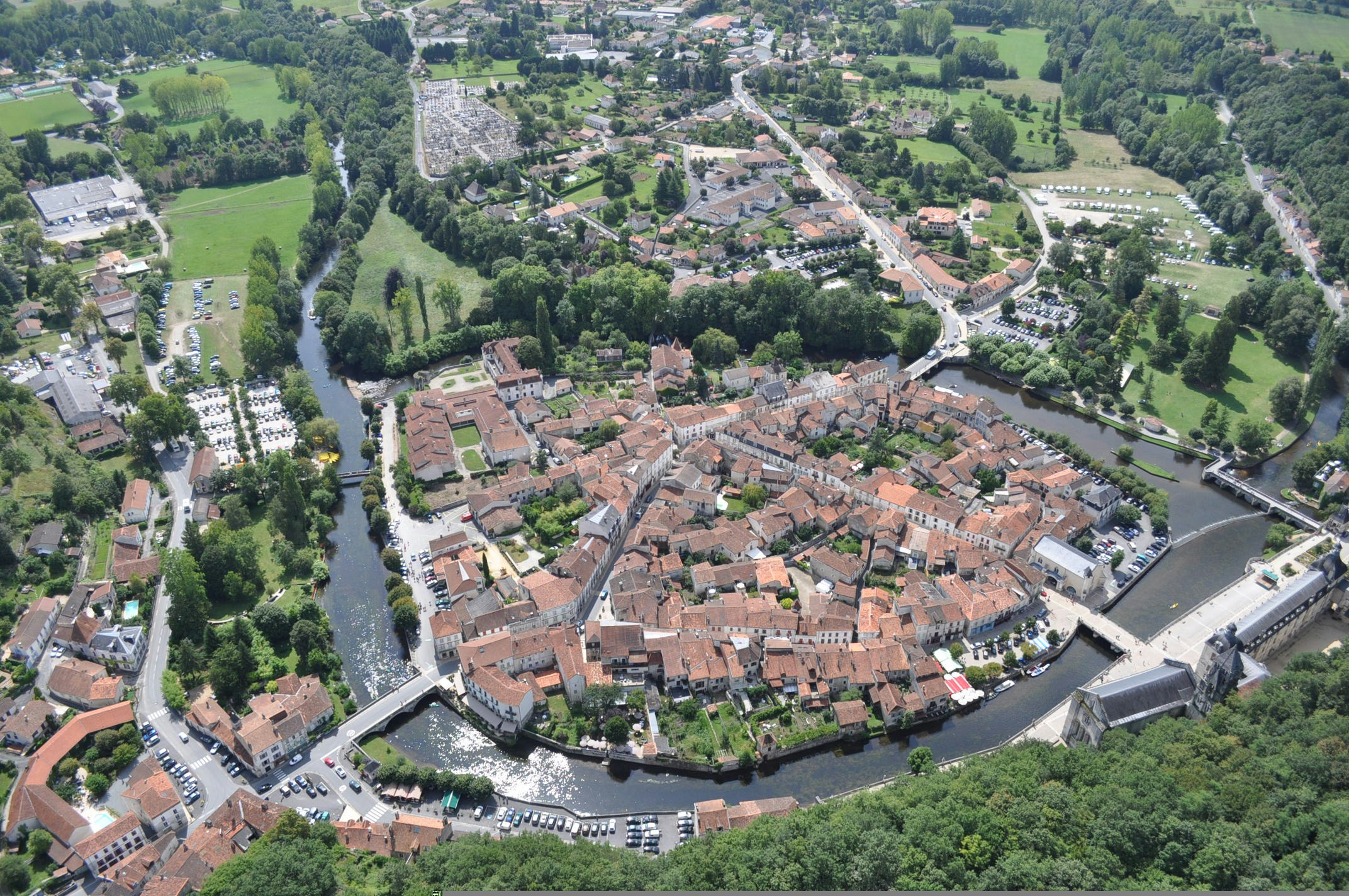 Luftaufnahme einer Altstadt mit Fluss, Brücken und Straßen, umgeben von grünen Wiesen und Bäumen im Tal.