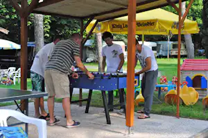 Tischkicker unter einem Holzpavillon auf dem Campingplatz, im Hintergrund gelbe Sonnenschirme und Spielgeräte für Kinder.