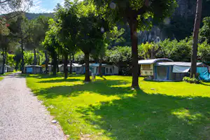 Campingplatz mit mehreren Wohnwagen und Mobilheimen zwischen hohen Bäumen, Grasfläche und Kiesweg am Rand.