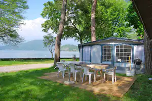 Campingunterkunft mit Terrasse am See, Tisch und Stühle unter Markise, umgeben von Bäumen und grünem Rasen.