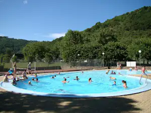 Kinder und Erwachsene im Camping-Schwimmbecken, umgeben von Bäumen und einem Zaun, im Hintergrund ein grüner Hügel.