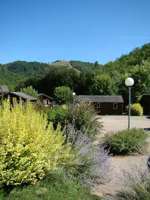 Campinganlage mit gepflegtem Garten, Kiesweg und mehreren kleinen Häusern, im Hintergrund bewaldete Hügel und Bäume.