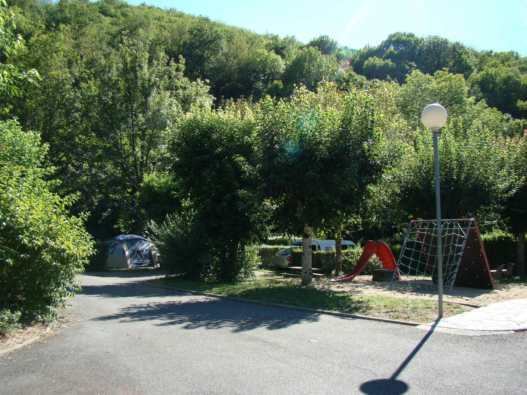 Spielplatz mit Rutsche und Klettergerüst auf einem Campingplatz, umgeben von Bäumen und Hecken.