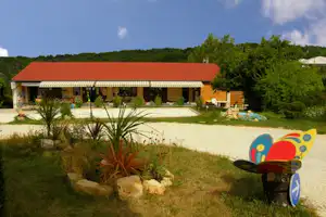 Bungalow mit rotem Dach und Terrasse, davor Kiesweg und Garten mit Sträuchern, Spielgerät im Vordergrund.