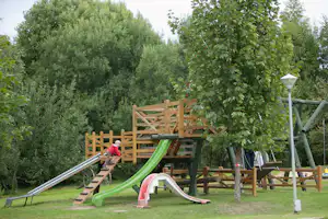 Spielplatz mit Klettergerüst und grünem Rutschenteil, umgeben von Bäumen und Rasenfläche auf dem Campinggelände.