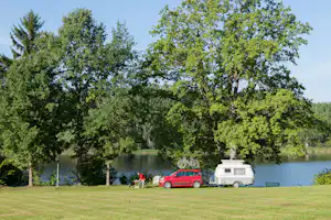 Campingplatz am See: rotes Auto und Wohnwagen am Ufer, umgeben von hohen Bäumen und grünem Rasen.