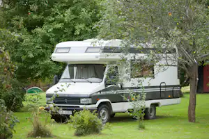 Weißes Wohnmobil auf einem grünen Campingplatz, von Bäumen und Sträuchern verdeckt, mit Front und Aufbaudach im Blick.