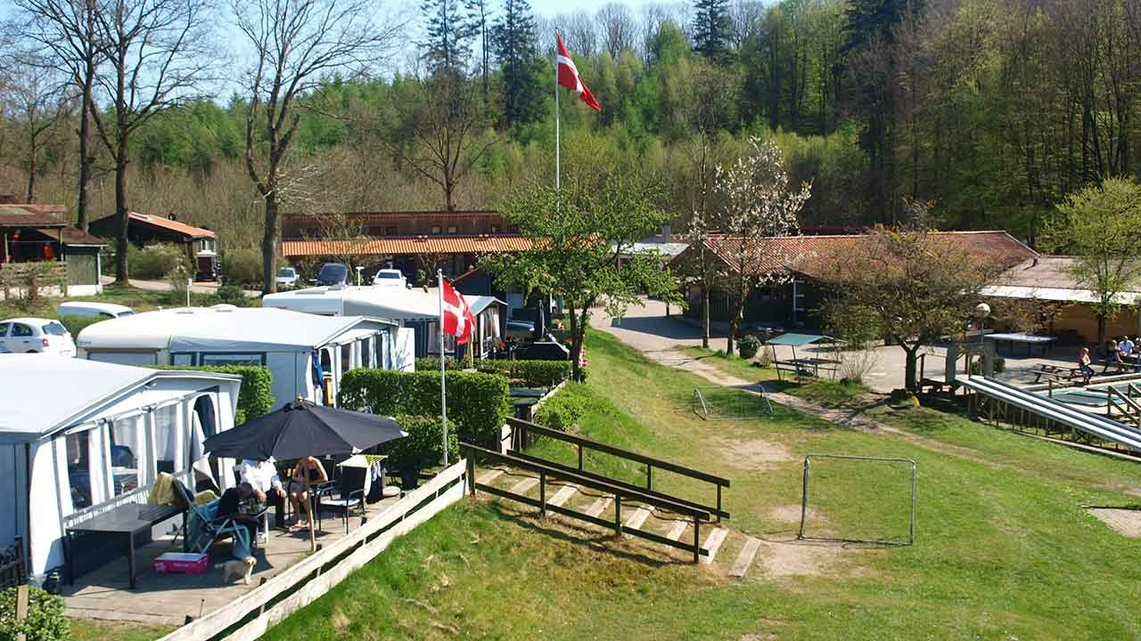 Reihe von Mobilheimen auf dem Campingplatz, mit Terrasse, Gartenzaun und Flaggen vor Bäumen und Wegen.