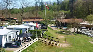 Reihe von Mobilheimen auf dem Campingplatz, mit Terrasse, Gartenzaun und Flaggen vor Bäumen und Wegen.