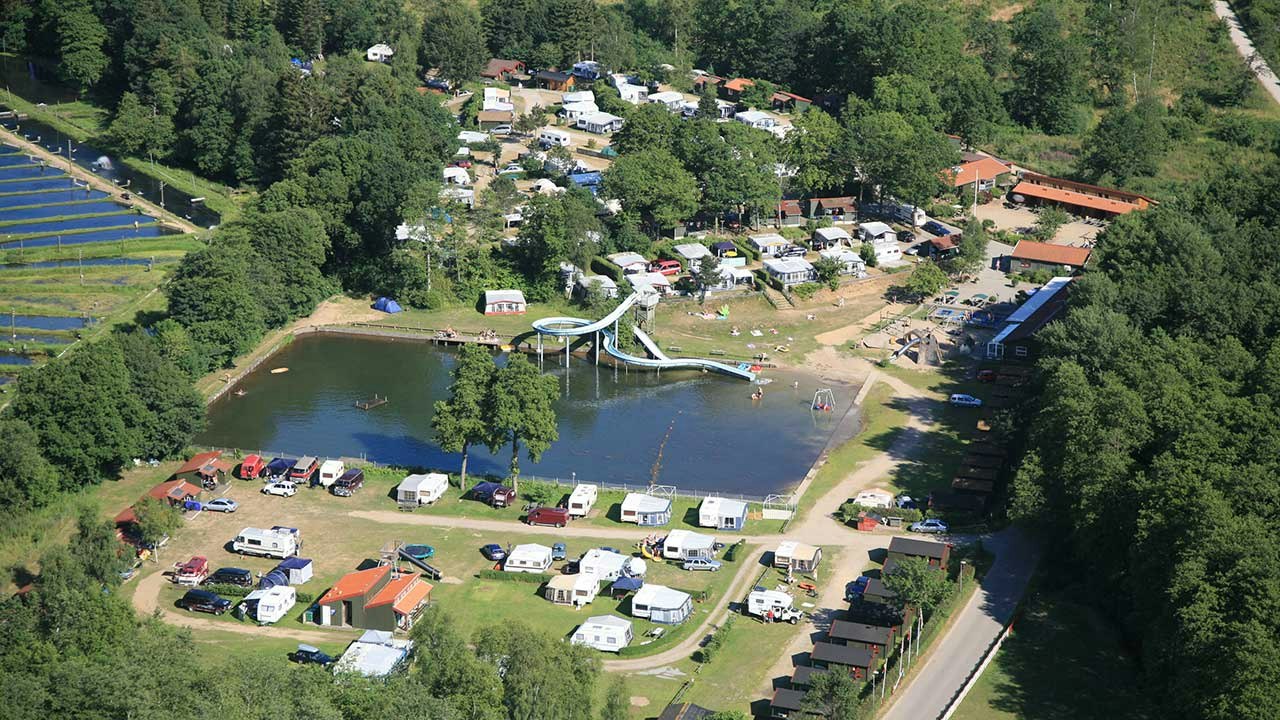 Luftaufnahme eines Campingplatzes am See mit Wohnwagen, Zelten, Wegen, Spielplatz und Bäumen im grünen Tal.