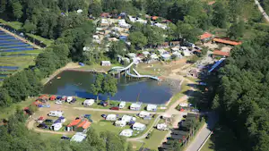 Luftaufnahme eines Campingplatzes am See mit Wohnwagen, Zelten, Wegen, Spielplatz und Bäumen im grünen Tal.