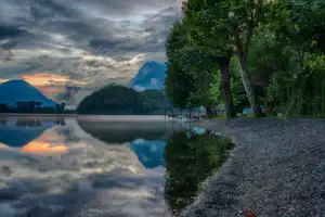 Seeufer mit Kiesstrand, hohen Bäumen und Holzpfählen im Wasser, dahinter Berge und Wolken am Abendhimmel.