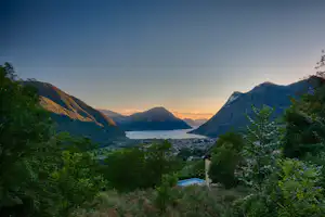 Blick ins Tal auf einen See zwischen Bergen, umgeben von Bäumen und einem kleinen Ort im Hintergrund.