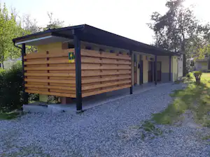 Camping-Unterkunft mit Holzverkleidung, überdachter Terrasse und Kiesweg, im Hintergrund Bäume und ein weiteres Gebäude.