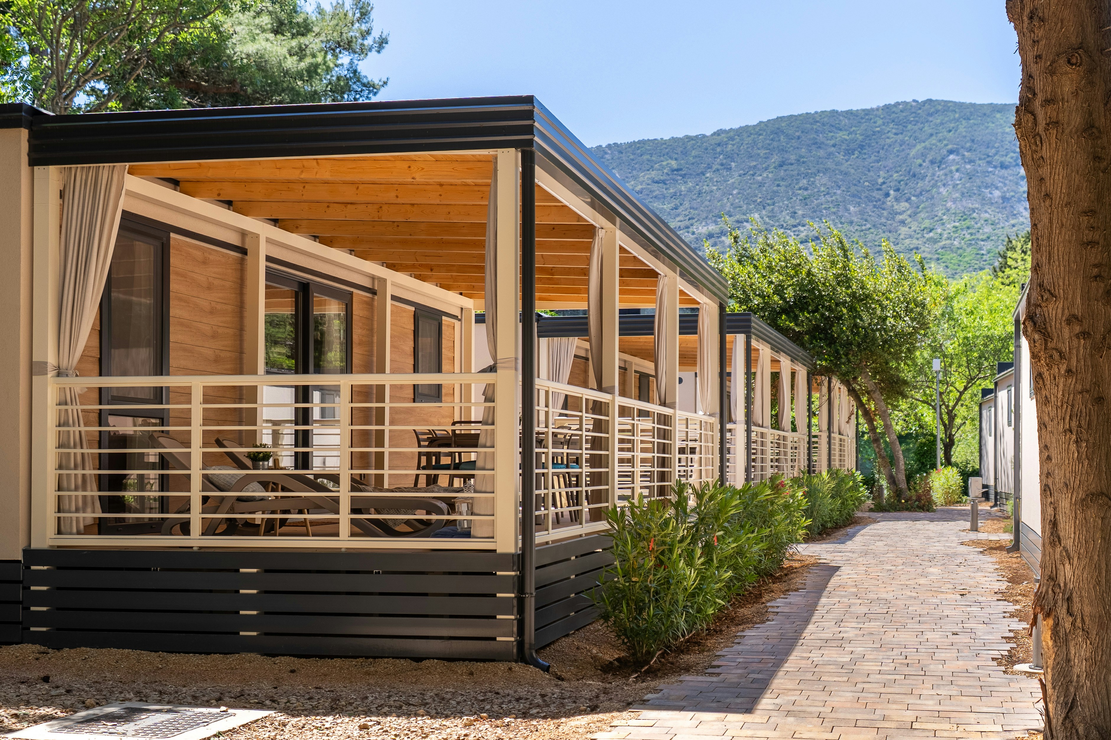 Reihen von modernen Ferienhäusern mit Holzveranda und Sichtschutz, gepflasterter Weg und Bäume vor den Bergen im Hintergrund.