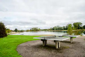 Picknicktisch am Campingplatz am See, mit Rasenfläche, Kiesweg und Bäumen am Ufer unter bewölktem Himmel.