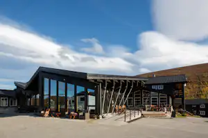 Moderner Campingbereich mit Glasfront, Sitzgruppe und Terrasse vor einem Gebäude, daneben Holzstapel und Wege auf dem Platz.