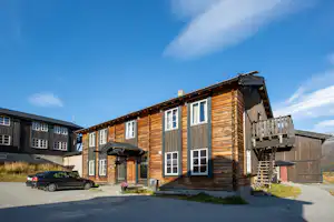 Holzhaus mit Balkon und Treppe, daneben ein Parkplatz mit Auto, im Hintergrund weitere Gebäude auf dem Campinggelände.