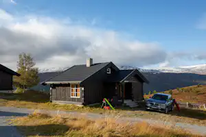 Holzferienhaus auf einem Campingplatz mit Terrasse, Auto vor dem Eingang und Bergen im Hintergrund.