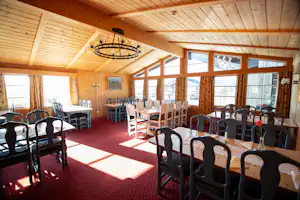 Speisesaal im Campinghaus mit Holzbalkendecke, vielen Stühlen und Tischen, großen Fenstern und rotem Teppichboden.