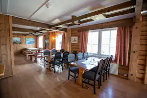 Essbereich in einem Holzhaus mit mehreren Tischen und Stühlen, großen Fenstern und Vorhängen, sichtbaren Holzbalken an der Decke.