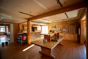 Innenraum eines Holzhauses mit Küchenzeile, Esstisch und Sitzbank, Regalen und Holzdecken, viel Tageslicht.