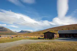 Holzhäuser und Wohnwagen auf einem Campingplatz am Weg, mit Wiesen, Bergen und blauem Himmel im Hintergrund.