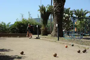 Mann läuft auf einem Campingplatzweg, im Vordergrund Boule-Kugeln, daneben Palmen und ein Metallgeländer an einer Steinmauer.
