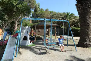 Spielplatz mit blauer Rutsche und Schaukeln, Sandboden und Holzhütte im Hintergrund zwischen Bäumen und Palmen.