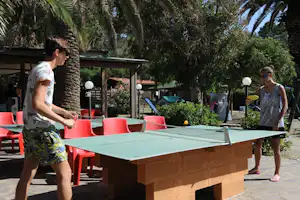 Zwei Personen am Tisch mit roter Campingmöblierung auf einer Terrasse zwischen Palmen und Büschen.