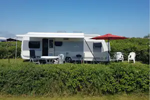 Wohnwagen mit Terrasse, Markise und rotem Sonnenschirm auf einem Campingplatz, umgeben von Hecken und Rasen.