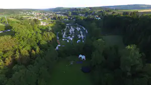 Luftaufnahme eines Campingplatzes im Tal mit vielen Stellplätzen, kleinen Gebäuden und Wegen zwischen Wäldern und Hügeln.