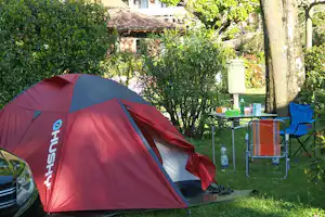 Rotes Campingzelt im grünen Garten mit Tisch, Stühlen und Wasserflasche neben dem Zelt.