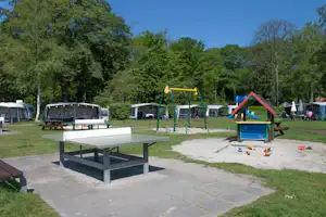 Tischtennisplatte auf dem Campingplatz, daneben Spielplatz mit Rutsche und Schaukel, im Hintergrund Zelte und Bäume.
