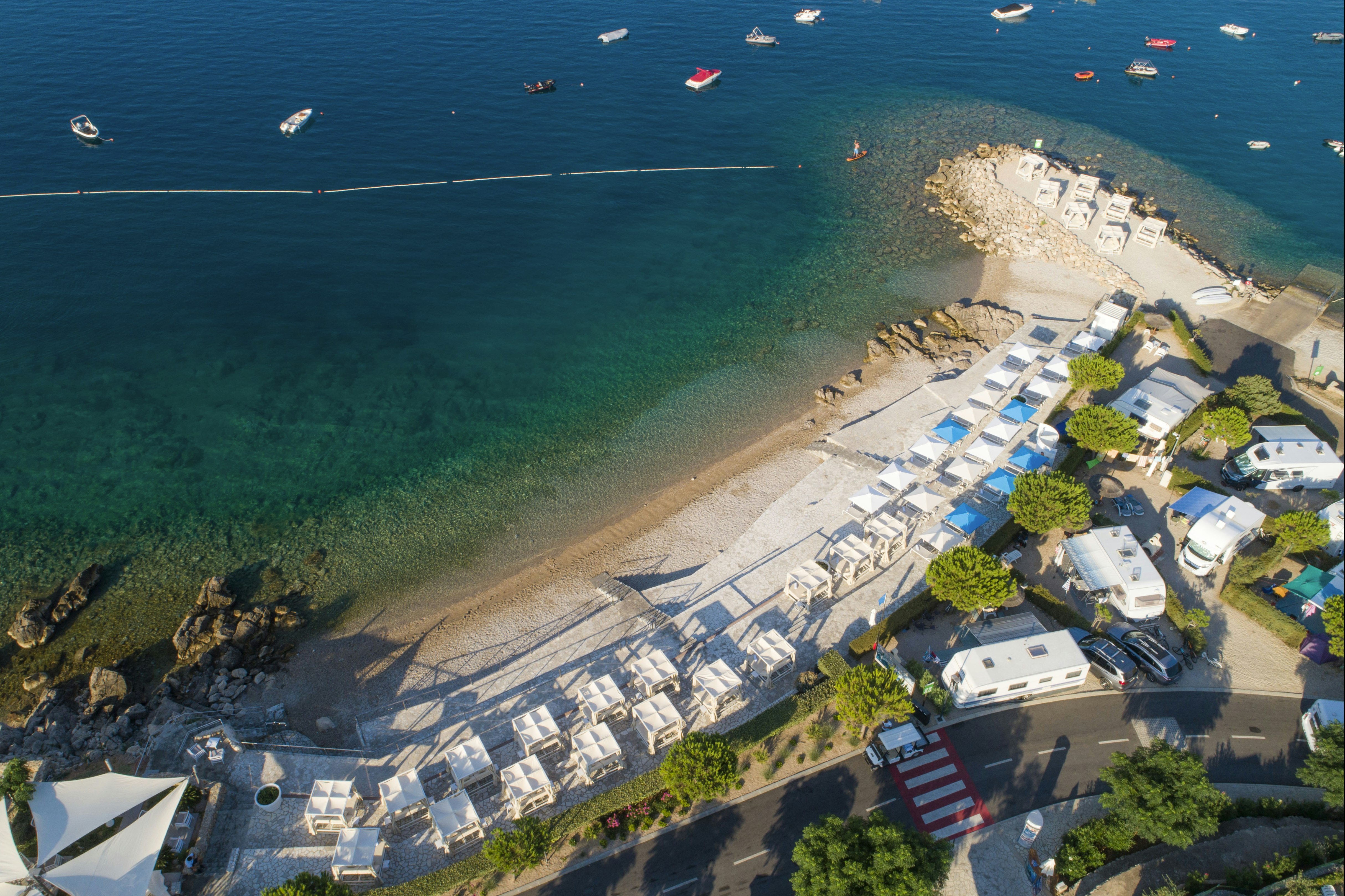 Campingplatz am Meer mit Strand, Steg und vielen Booten im klaren Wasser, daneben Stellplätze mit Wohnwagen.