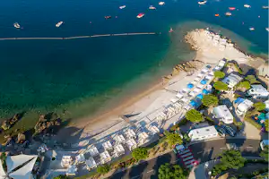 Campingplatz am Meer mit Strand, Steg und vielen Booten im klaren Wasser, daneben Stellplätze mit Wohnwagen.