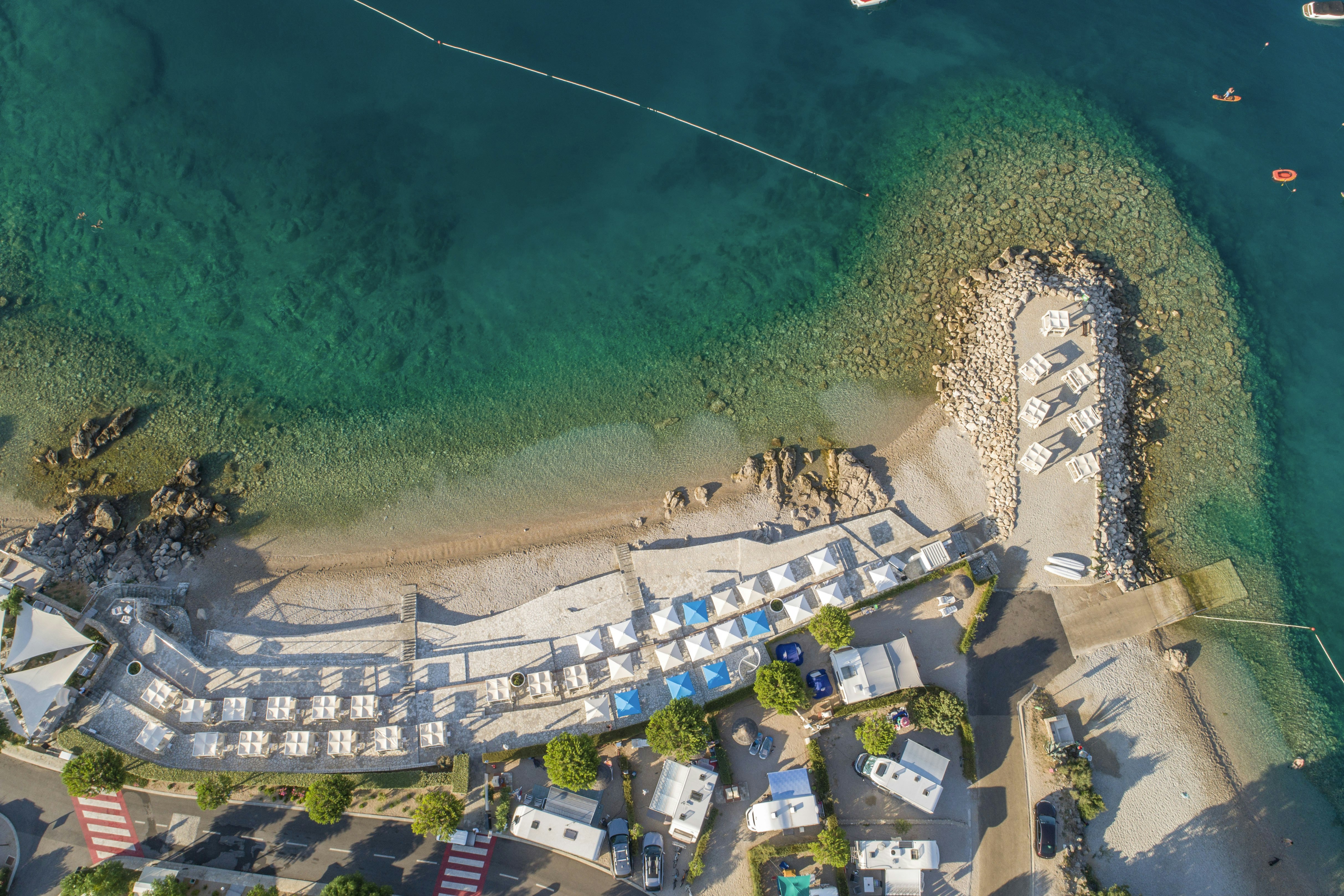 Campingplatz am See mit Strand, Liegen und Poolbereich, umgeben von Wegen, Bäumen und Gebäuden am Ufer.