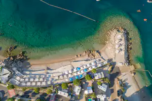 Campingplatz am See mit Strand, Liegen und Poolbereich, umgeben von Wegen, Bäumen und Gebäuden am Ufer.